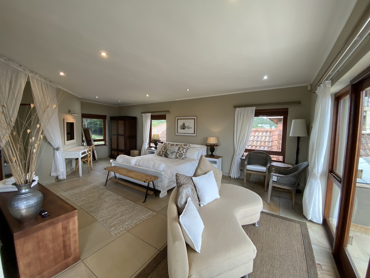 A bright bedroom with white bedding, chaise and armchairs by the windows